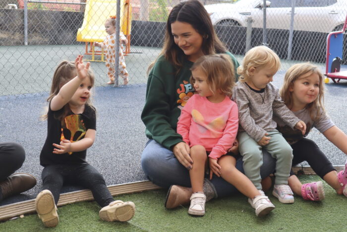 A casually dressed young woman sits with several young children as one raises her hand to answer a question.
