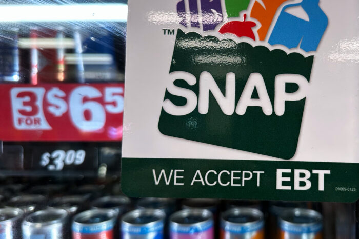 A sticker with SNAP branding declares "We accept EBT" on the glass front to a refrigerator containing rows of canned drinks. A partial price of $6, printed in large white characters on a red background can be seen behind the sticker.