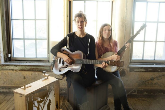 Two white women standing in a room in front of a window. The woman on the left is holding a guitar, and the woman on the right is holding a banjo. 