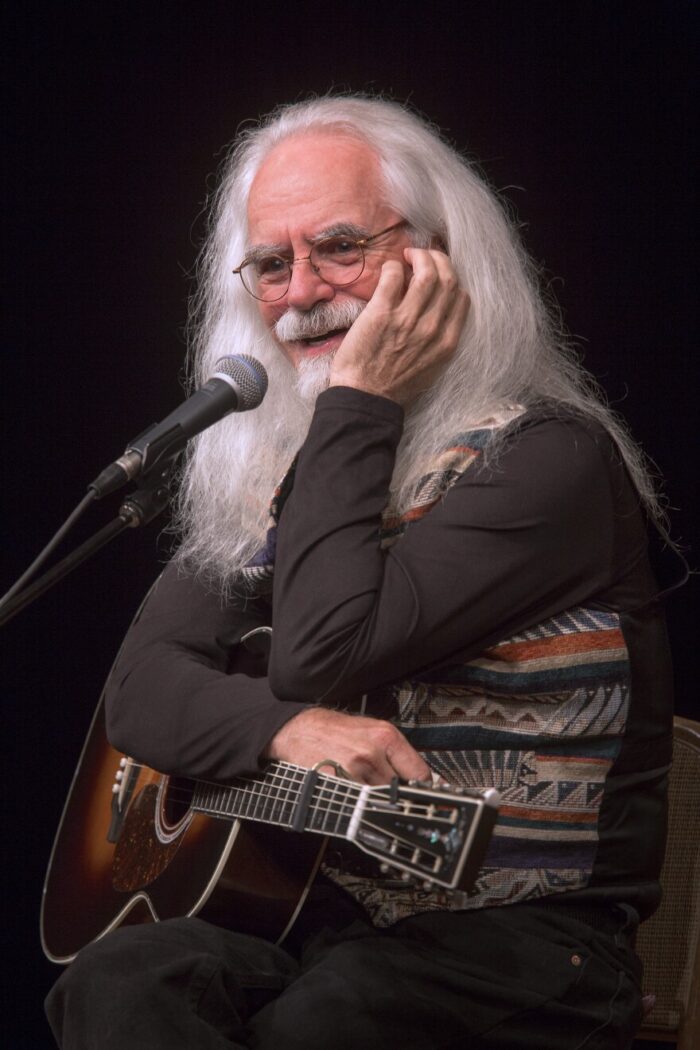 A white man sitting on a stool in front of a microphone. He is resting his arms on a guitar. One hand is against his face.