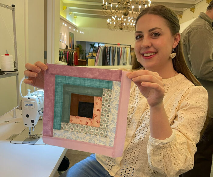 A smiling young woman holds up a quilted square made with multiple thin, long rectangular scraps of material.