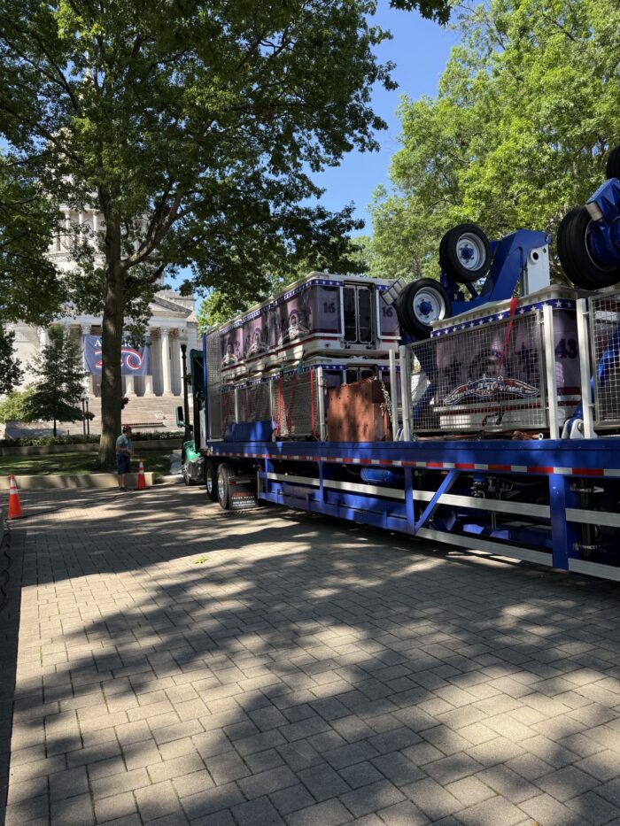 Navy blue gondolas sit on an 18-wheeler bed beneath the tree-shaded West Virginia Capitol.