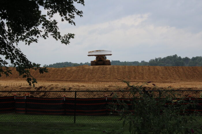 A bulldozer with earth moving blade lifted crests a bare, earthen dome. The heavy machinery looms over a verdant green yard.