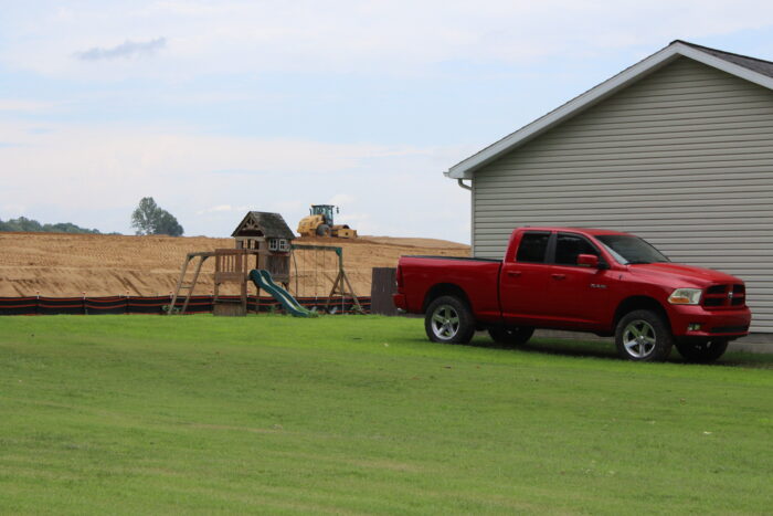 A red pickup truck is parked next to a home with a play set in the backyard. Immediately behind the playset can be seen heavy machinery on a high berm of bare dirt with a clouded sky above.