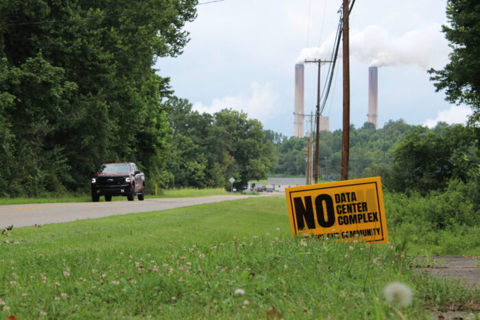 A yellow lawn sign reads " No Data Center Complex" in black lettering. The sign sits under a row of power lines leading into the background towards smoke stacks that protrude into the sky above trees. A black pickup drives past the sign on a road to the left.