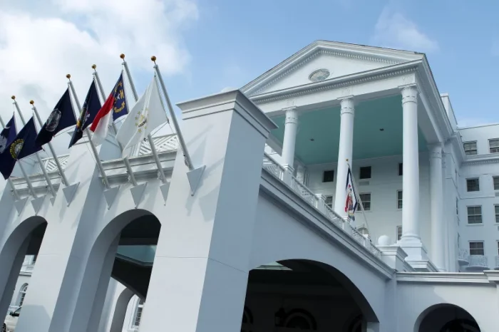 A white building with several flags flying out front. The building features white pillars and is seen from a low angle against the blue sky as a backdrop.