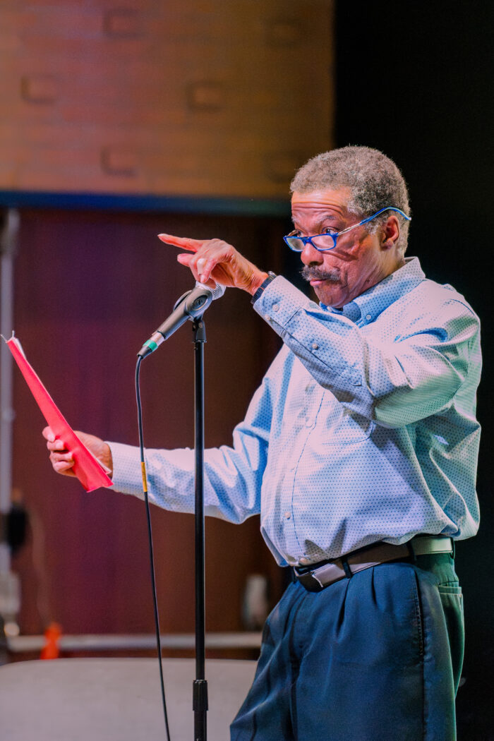 A Black man stands at a standing mic holding a red piece of paper. He wears glasses and is dressed in business casual. He points toward the panelists off camera.