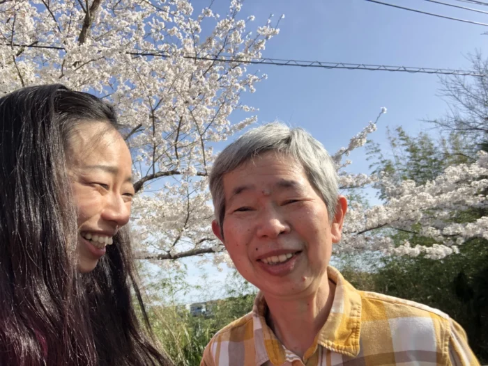 Two Japanese women smiling at the camera. They are outside and there is a cherry blossom tree behind them. 