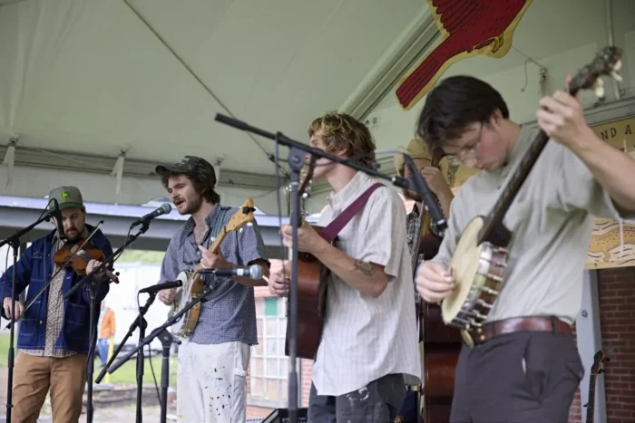 Four young white men standing on a stage and singing into a microphone. They are all holding string instruments. 