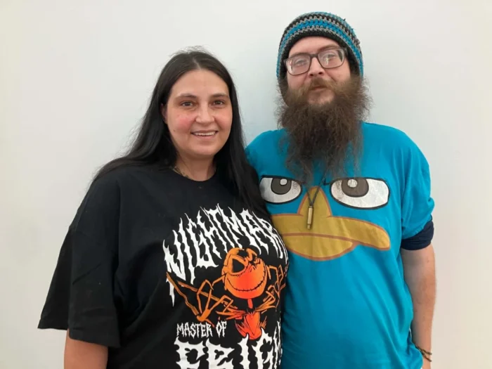 A white woman and man people posing for a photo. One is wearing a black shirt and the other a blue shirt. 