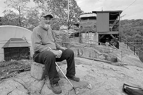 A black-and-white photo of a man sitting on a rock. There is a bus behind him.