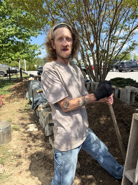 A white man holding a shovel on a pile of dirt. He is standing outside and wearing a gray shirt and blue jeans. 
