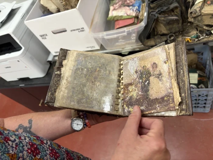Two hands holding a damaged photo album. The person holding the photo album is standing above a table with other items on top of it. 