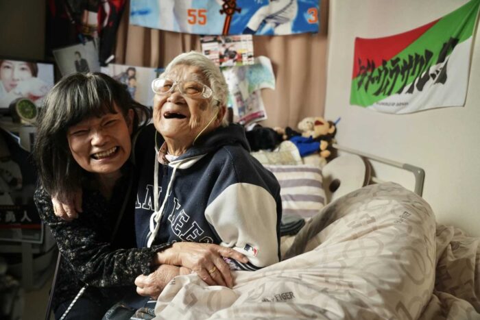 Two older Japanese women smiling. They are sitting on a bed and hugging. 