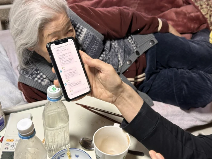 A photo of a woman holding a phone. There is an elder Japanese woman lying in front of her. 