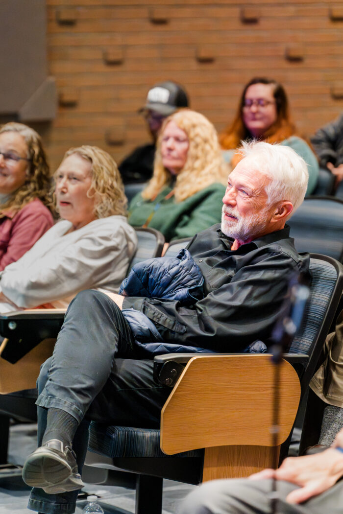 A white man with white hair sits comfortably in a chair in an auditorium. He looks on toward the stage. Behind him are other seated people attending the event.