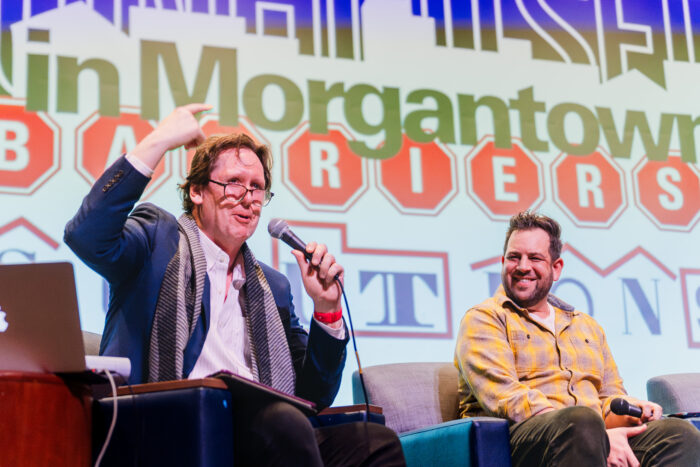 Two men are seated on a stage. The man on the left is Us & Them host Trey Kay. He is speaking into a microphone and gesturing toward the audience.
