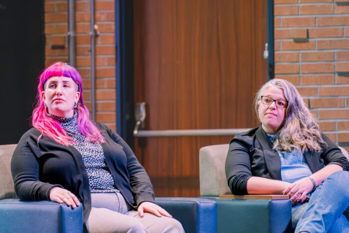 Two women sit next to each other on a stage. The woman on the left has pink hair, while the woman on the right has grayish hair. They both appear to be listening to someone speaking.