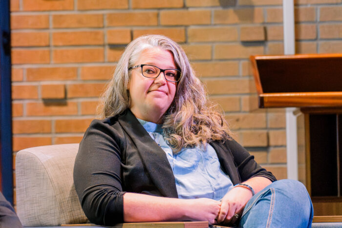 An older woman sits in a chair on a stage. She wears a black blazer, glasses, and jeans.