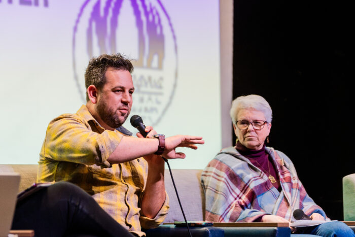 Two panelists, a man and a woman, are seated on a stage. The man is holding a microphone and is mid-sentence. He is gesturing with his hand. The woman looks on, listening.