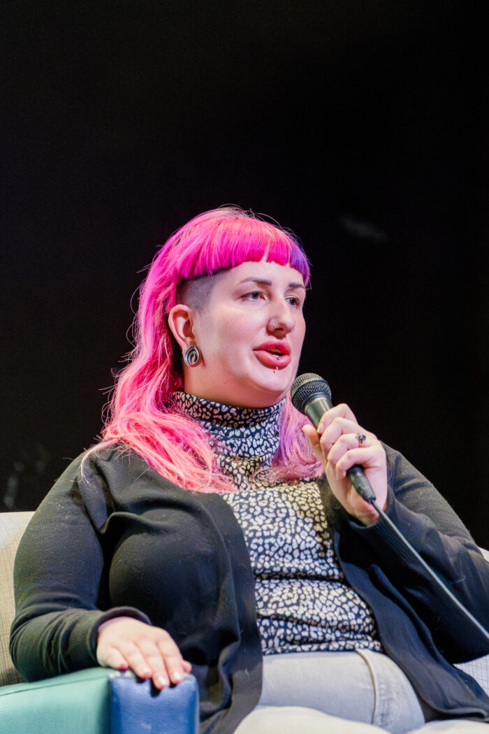 A woman with pink hair and facial piercings speaks into a microphone as she sits in a chair on a stage. She is wearing a black sweater.