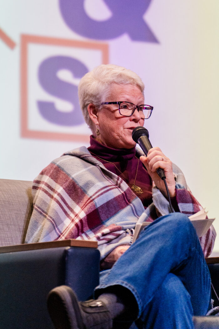A close up of an older woman wearing glasses. She is also wearing a plaid poncho.