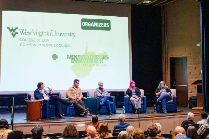 Five people sit on a stage in chair. They are speaking before an audience. Behind them is a projector screen with text. There are several people in the audience with their backs to the camera.