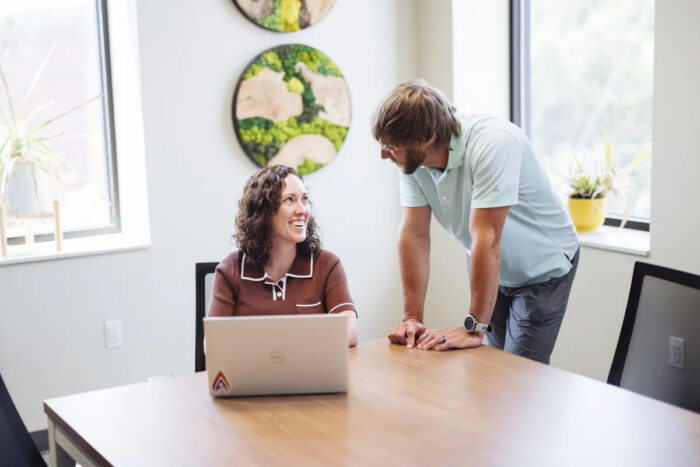 Woman sits at a table with computer while man with beard in blue shirt looks at her.