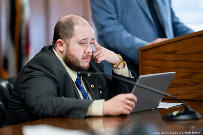 A balding man with a brown bears sits at a desk and looks at the screen of a laptop. He is formally dressed with a black blazer and blue tie.