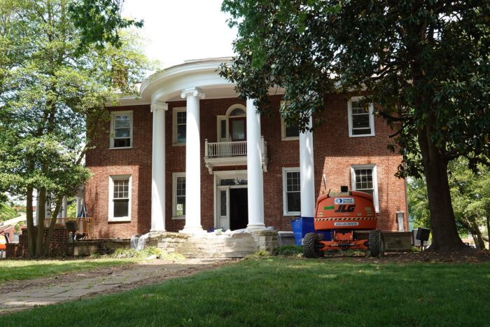 Photo shows a two-story, red brick home with large white columns forming a circular arch around the front porch. To either side of the porch is construction machinery.