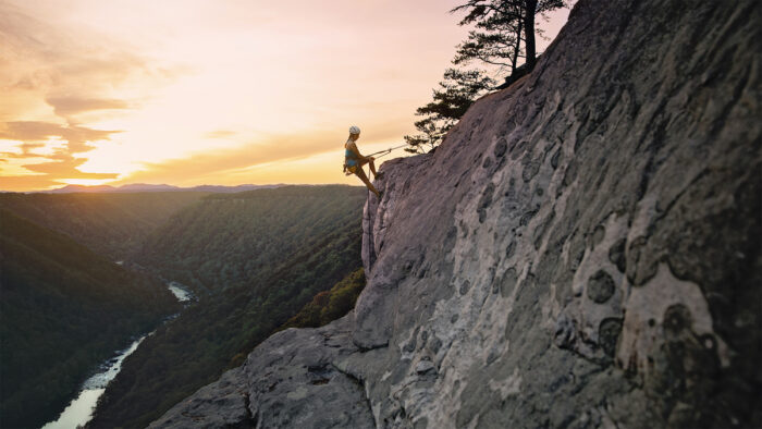 Photo shows silhouette of a young woman climbing rocks above a scenic river with a golden sunset in the distance.