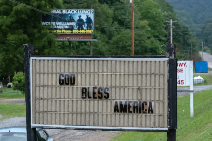 A billboard seen against a hill of green trees. Words on the billboard read, "God Bless America."
