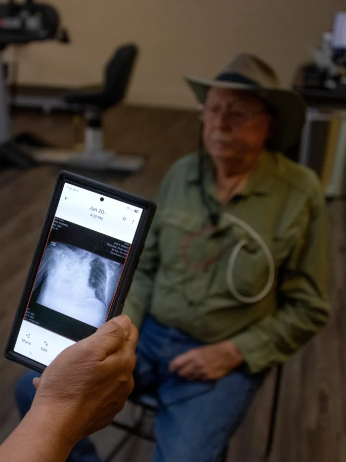 An older man with white hair, wearing a cowboy hat, sits in a chair looking off camera. Someone hand is visible just to the side of the photo. The hand holds a tablet looking at a photo of someone with black lung disease.
