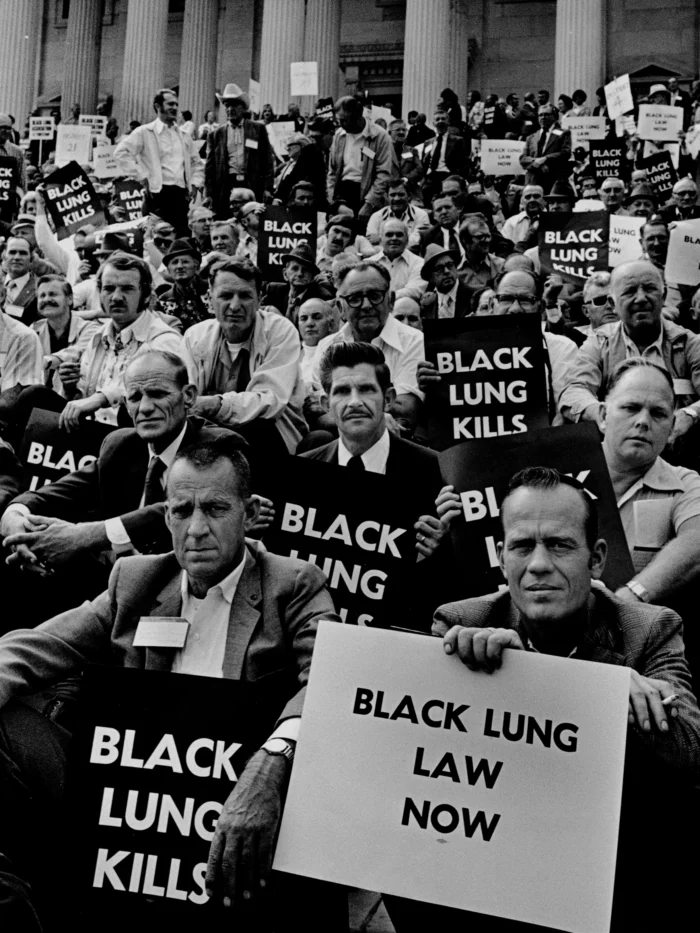 A black and white photo of dozens of people sitting on the steps of the U.S. Capitol. They are holding signs that read, "Black Lung Kills," and "Black Lung Law Now."