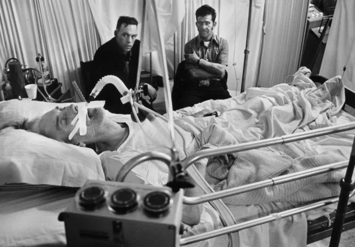 A black and white photo of a man dying in a hospital bed. He has a tub down his throat to help him breath. Two men sit next to him, looking toward the camera, not smiling.