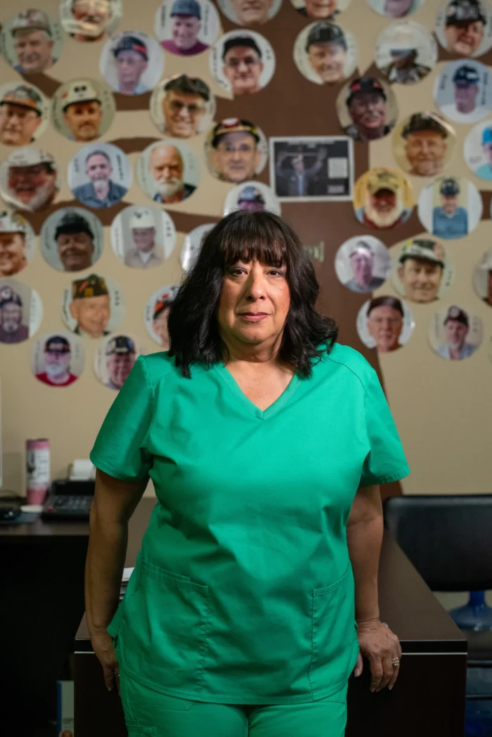 A woman dressed in medical scrubs with dark hair looks toward the camera, not smiling. Behind her is a wall of photos.