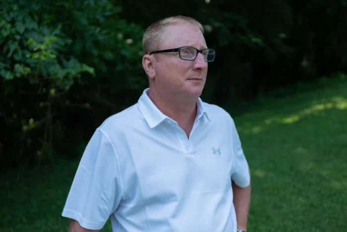 A close up of an adult man wearing glasses and with very short hair. He looks off camera with his hands on his hips.