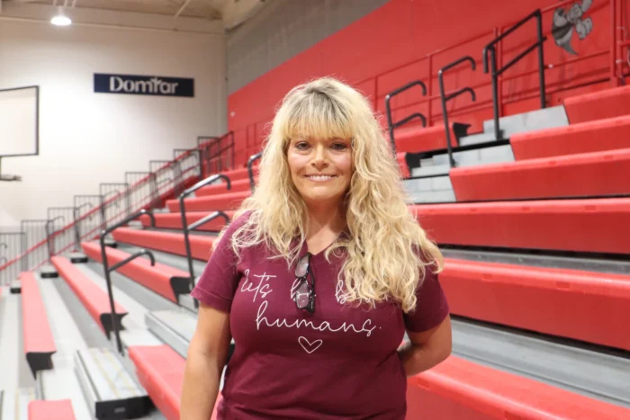 An adult woman with long blond hair smiles for the camera. She is wearing a maroon shirt with text that reads, Let's Be Better Humans. Behind her are red bleachers inside a gymnasium.
