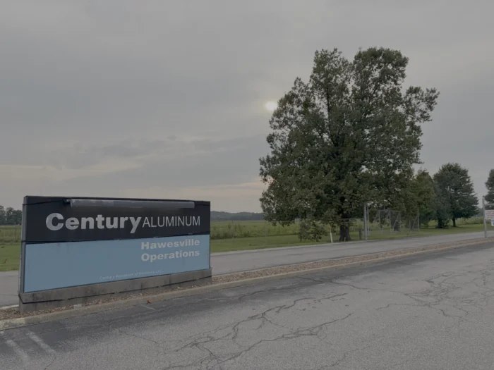 Outside on a cloudy day. A sign on a concrete road reads, Century Aluminum, Hawesville Operations. In the distance are trees and a field.