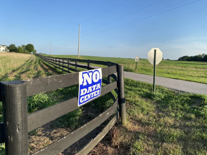 A wooden fence, on a clear, sunny day, is shown wrapped around a farm on several aches of land. A sign reads on the sign, "No Data Center."