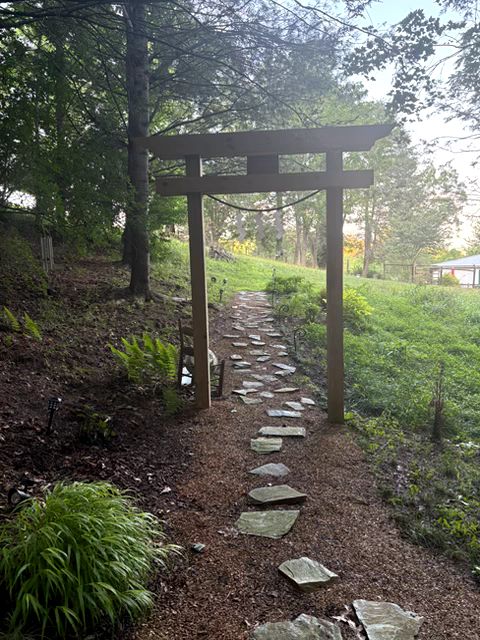 A wooden entrance to a garden. There are stones on the forest floor leading to the garden's entrance.