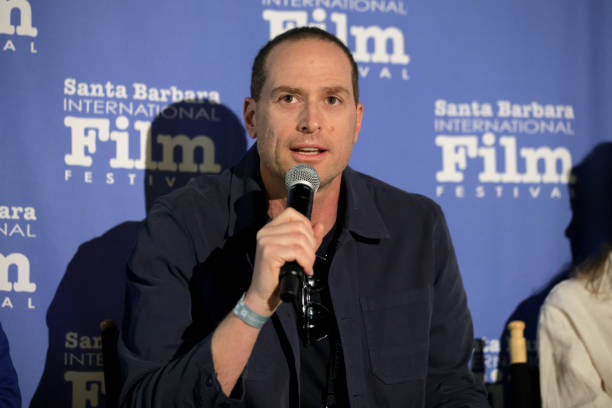 A professional photograph of an adult man with shortly trimmed hair. He speaks into a microphone and looks off camera. He wears a brown jacket and stands in front of a poster with text that reads, Santa Barbara International Film Festival.
