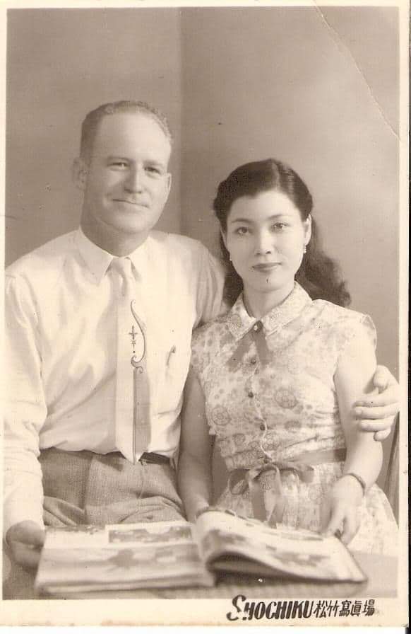 A black-and-white portrait of two people. On the left, a white man wraps his arm around the Japanese woman on the right. There is a book laying out in front of them.