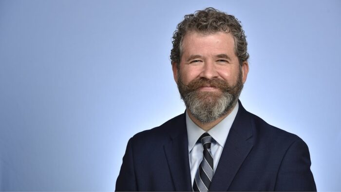 A professional photograph of an adult man with salt and pepper hair, a beard, and a mustache that has been curled at the ends. He is formally dressed in a blue suit and tie.