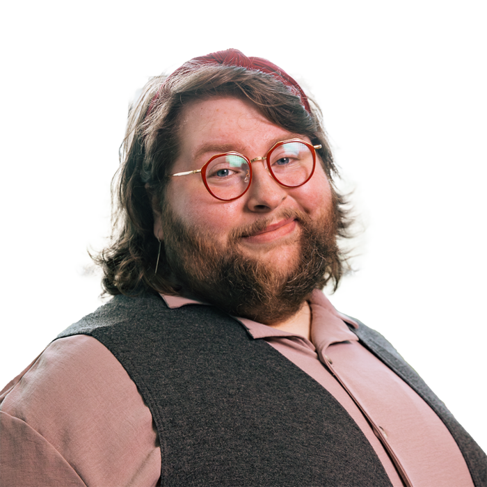 A professional photo of a bigger man with brown hair and a brown beard. He wears red-rimmed glasses, a gray shirt, and a dark vest over it.