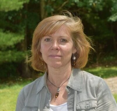 A woman looks toward the camera, not smiling. She has light red hair and wears earrings. She has a gray cardigan on over a white shirt. She is outside.