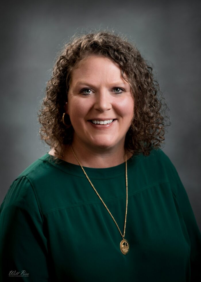 A professional photo of a woman wearing a green shirt and golden necklace. She has short, curly brown hair.