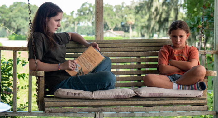 Two girls, one older and one younger, sit on a porch swing. The younger girl has her arms folded across her chest and looks down, frowning. The older girl sits looking toward her, also looking concerned, but is looking at her.