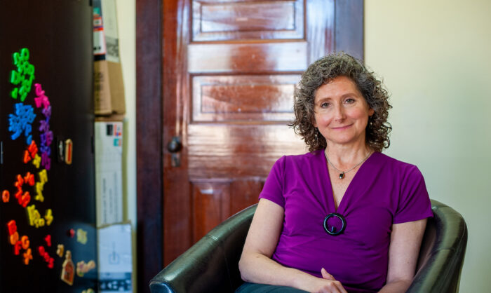 An older woman sits in a pleather chair and softly smiles toward the camera. She has salt and pepper, curly hair and wears a purple shirt.