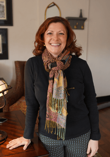 An adult woman with dark red hair smiles and looks toward the camera. She is wearing a black sweater and a colorful scarf.